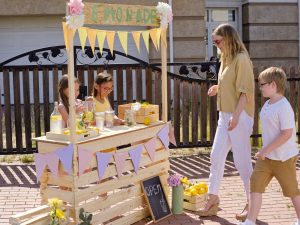 Young elegant blond woman and her little son coming up to wooden stall where two cute girls selling fresh cool homemade lemonade